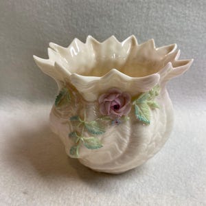May include: Cream ceramic vase with a scalloped rim and a ribbed body. A pink rose and green leaves with blue accents decorate the front. The vase is a decorative item.