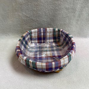 May include: A round, clear plastic serving dish with three divided sections, nestled inside a fabric basket liner featuring a plaid pattern of white, blue, green, and red. The liner has a gathered edge that wraps around the exterior rim.