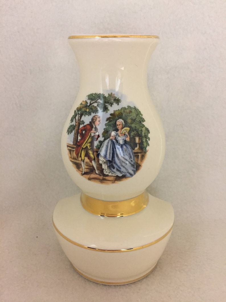 May include: A white ceramic vase with a gold rim and base. The vase features a painted scene of a man and woman in a garden setting.