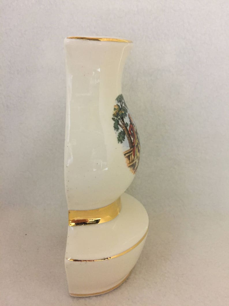 May include: A white ceramic vase with a gold trim and a painted scene of a couple in a garden. The vase is shaped like a traditional pitcher with a wide base and a narrow neck.