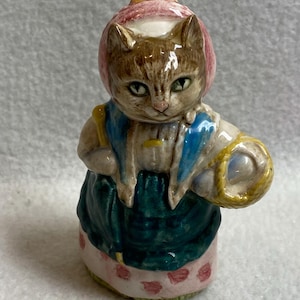 May include: A ceramic figurine of a cat dressed in a bonnet and apron. The cat has brown fur, green eyes, and is wearing a pink bonnet, blue apron, and a green dress. It is holding a yellow basket.