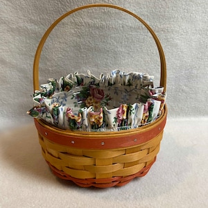 May include: A small, woven basket with a natural wood handle and a floral fabric liner. The basket is brown and orange with a white and floral patterned liner.