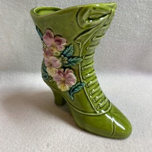 May include: A green ceramic boot-shaped vase with a high heel and floral accents. The vase is a light green color with pink and yellow flowers and green leaves. The boot has a lace-up design.