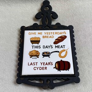 May include: A white ceramic trivet with a black cast iron frame. The trivet features a humorous saying: "Give me yesterday's bread, this day's meat, and last year's cyder". The saying is accompanied by illustrations of bread, meat, and a barrel of cider.