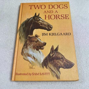 Puede incluir: Un libro antiguo titulado "TWO DOGS AND A HORSE" de Jim Kjelgaard. La portada presenta ilustraciones de un caballo y dos perros en tonos marrones y negros. El libro está ilustrado por Sam Savitt.