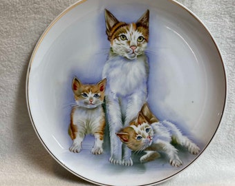 May include: A white ceramic plate with a gold rim features a painted image of a white cat with brown and orange markings and two white kittens with brown and orange markings. The cat and kittens are looking at the viewer.