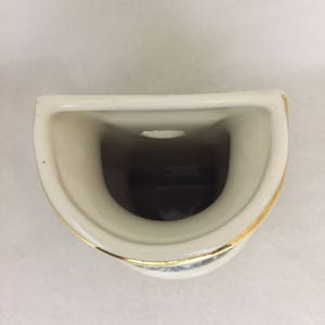 May include: A white ceramic toothbrush holder with a gold rim. The holder has a single opening for a toothbrush.