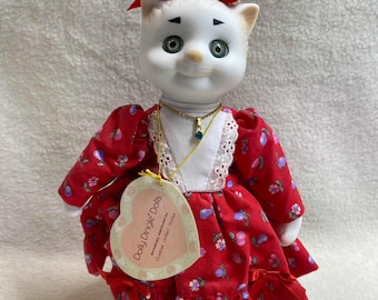 May include: A white porcelain cat doll wearing a red dress with white flowers and a gold necklace with a blue gem. The doll has green eyes and a red bow with a white flower on its head. The doll is labeled "Dolly Dingle Dolls" with the text "exclusively manufactured by Golden Living Stores".