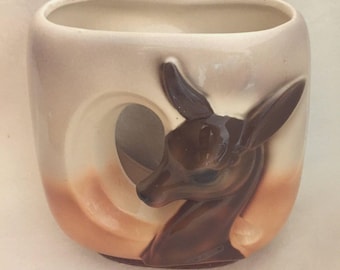May include: A ceramic vase with a deer design. The vase is rectangular with a curved top and a circular cutout. The deer is brown with a white and tan background. The vase is tan and brown.