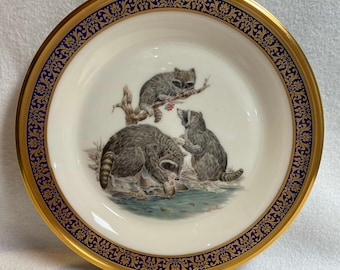 May include: A white porcelain plate with a gold rim and blue and gold floral border. The center of the plate features a painting of three raccoons in a woodland setting.
