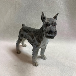 May include: A gray and white miniature Schnauzer figurine. The dog is standing with its head up, ears perked, and a detailed, textured coat. The figurine has a realistic appearance, with a focus on the dog's facial features and body posture.