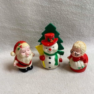 May include: A set of three Christmas figurines: Santa Claus, a snowman with a red hat and green scarf, and Mrs. Claus. The figurines are painted with bright colors and have a vintage aesthetic.