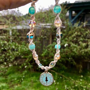 May include: A white macrame cord necklace with turquoise and gold beads and a gold charm with a clock face and a tree silhouette.