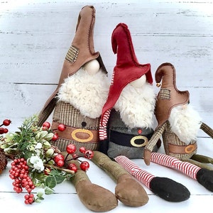 Może przedstawiać: Trzy ręcznie robione lalki krasnoludków z czerwonej, brązowej i białej tkaniny. Krasnoludki siedzą na białym drewnianym podłożu z zielonymi i czerwonymi jagodami.