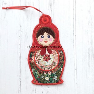 May include: Red and green fabric Christmas ornament shaped like a Russian nesting doll with a floral pattern and a smiling face. The ornament has a loop for hanging and the text "Pixie Willow Patterns" is visible.