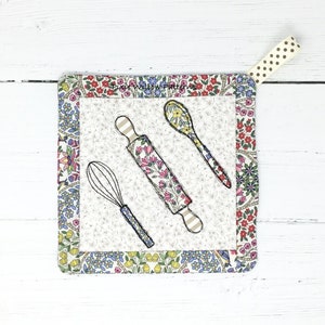 May include: A white cotton pot holder with a floral print border. The centre features a black and white illustration of a whisk, rolling pin, and spoon.
