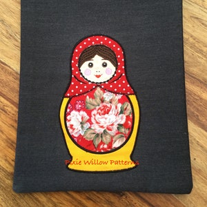 May include: A grey fabric pouch featuring an embroidered Russian nesting doll design in red, white and yellow. The doll has a floral pattern on its body and the text "Pixie Willow Patterns" is embroidered below the doll.