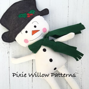 May include: A white snowman doll with a black top hat, green scarf, and black buttons. The snowman has a carrot nose, black eyes, and pink cheeks. The doll is lying on a white wooden surface. "Pixie Willow Patterns" is written in black text on the surface.