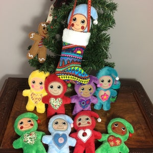 May include: A collection of eight plush toys, each with a different color and design. The toys are all wearing a heart-shaped patch on their chest. The toys are arranged in a circle around a Christmas stocking with a colorful pattern. The stocking is hanging from a Christmas tree.
