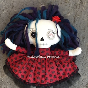 Könnte beinhalten: Handgemachte Puppe mit Totenkopfgesicht, blau-violettem Garnhaar und einer roten Rose. Die Puppe trägt ein rotes Kleid mit schwarzem Schneckenmuster und schwarzem Spitzensaum. Die Beine der Puppe sind grau und schwarz.