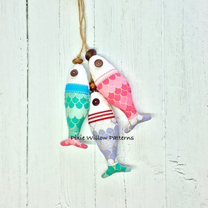 Könnte beinhalten: Drei Stofffisch-Dekorationen, die an einer Juteschnur hängen. Die Fische sind in Rosa-, Türkis- und Lavendeltönen gehalten, mit goldenen Schuppenmustern und Knopfaugen. Der Text "Pixie Willow Patterns" ist sichtbar.