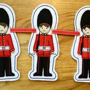 Könnte beinhalten: Ein rotes, weißes und schwarzes Filzbanner mit drei gestickten Bildern von britischen Gardisten in roten Uniformen, schwarzen Hüten und Gewehren. Das Banner ist mit einem roten Band mit weißen Nähten verziert.