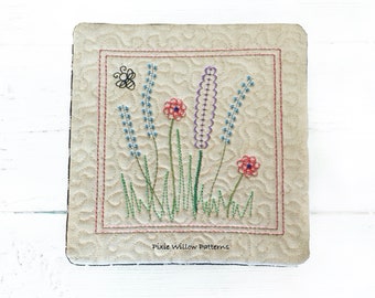 May include: A beige fabric square with a stitched border and a floral embroidery design. The design features purple and blue flowers, green leaves, and red flowers. The text "Pixie Willow Patterns" is stitched in the bottom center of the square.