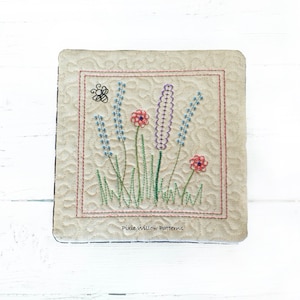 May include: A beige fabric square with a stitched border and a floral embroidery design. The design features purple and blue flowers, green leaves, and red flowers. The text "Pixie Willow Patterns" is stitched in the bottom center of the square.