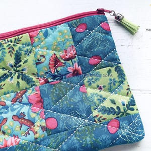 May include: A quilted zippered pouch with a patchwork design featuring floral and geometric patterns in shades of pink, green, and blue. The pouch has a light pink zipper and a green tassel.