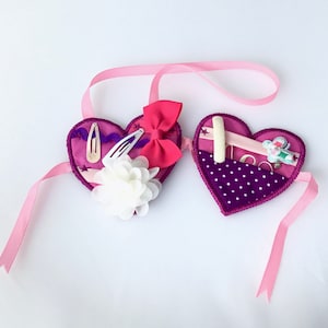 Pode incluir: Dois organizadores em forma de coração em tons de roxo, conectados por uma fita rosa. Um coração contém grampos de cabelo, um laço e uma flor branca. O outro coração tem um bolso com pequenos objetos. Um acessório charmoso.