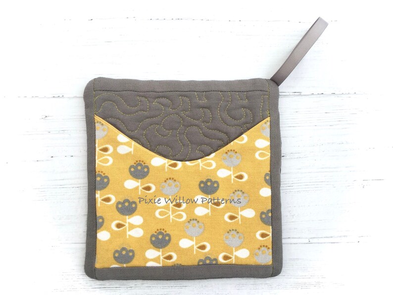May include: A gray and yellow quilted pot holder with a floral pattern and a gray loop handle. The pot holder has the text "Pixie Willow Patterns" printed on it.