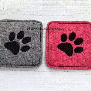 Puede incluir: Dos posavasos cuadrados de fieltro con bordado de huella de pata negra. El posavasos de la izquierda es gris y el de la derecha es rosa. El texto "Pixie Willow Patterns" est&aacute; bordado en los posavasos.