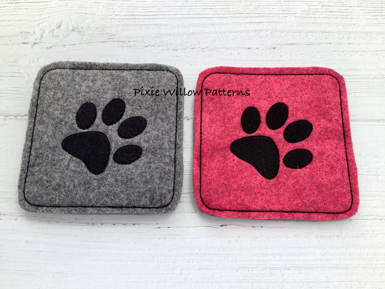 Puede incluir: Dos posavasos cuadrados de fieltro con bordado de huella de pata negra. El posavasos de la izquierda es gris y el de la derecha es rosa. El texto "Pixie Willow Patterns" est&aacute; bordado en los posavasos.
