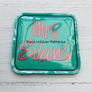May include: A teal blue square patch with a black border and the text "Mrs. Evans" in pink cursive lettering. The text "Pixie Willow Patterns" is also included in black lettering.