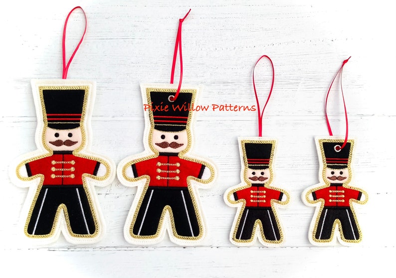 May include: Four felt Christmas ornaments in the shape of a gingerbread soldier. The ornaments are red, black, white, and gold. The ornaments have a red ribbon for hanging.