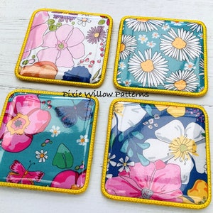 May include: Four square fabric coasters with a yellow stitched border. Each coaster features a different floral pattern with pink, white, and yellow flowers on a blue, green, or white background. The coasters are arranged in a square pattern on a white wooden surface.  "Pixie Willow Patterns" is written in black text on the surface.