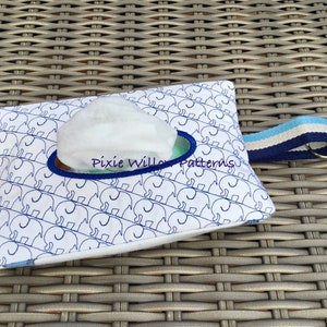 Pode incluir: Porta-lenços branco com estampa de elefantes azul e branco. O porta-lenços tem uma borda azul e uma alça azul com uma faixa branca e um chaveiro prateado. O texto "Pixie Willow Patterns" é bordado na frente do porta-lenços.