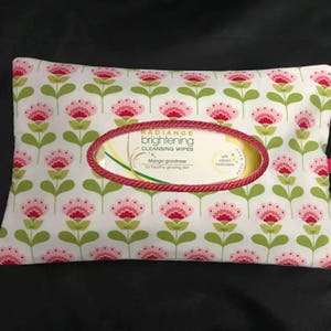 May include: A rectangular, floral-patterned wipe case with a pink ribbon pull. The case features a white background with a repeating pattern of pink flowers and green leaves. The center has an oval opening with a red rope trim, displaying the text "Radiance Brightening Cleansing Wipes".