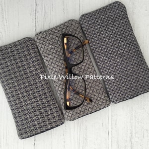 May include: Three gray and black patterned fabric eyeglass cases. The cases are lined up horizontally on a white wooden surface. The middle case has a pair of tortoiseshell glasses inside. The text "Pixie Willow Patterns" is visible on the middle case.