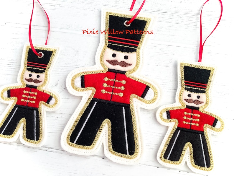 May include: Three felt Christmas ornaments shaped like gingerbread men, each wearing a red and black uniform with gold trim. The ornaments are decorated with gold buttons and a black hat with gold trim. The largest ornament is approximately 6 inches tall, the middle ornament is approximately 4 inches tall, and the smallest ornament is approximately 3 inches tall.