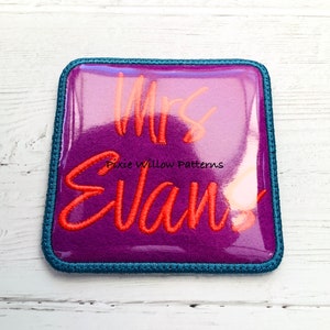 May include: A purple felt square with a blue border, featuring the text "Mrs. Evan!" in orange embroidery.