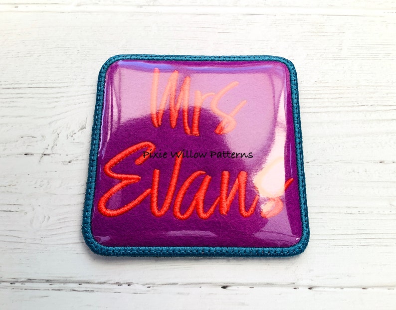 May include: A purple felt square with a blue border, featuring the text "Mrs. Evan!" in orange embroidery.