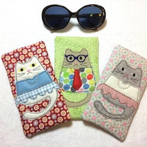 Puede incluir: Tres estuches para gafas de tela con diseños de gatos bordados. Los estuches son rojos con lunares blancos, verdes con remolinos blancos y grises con lunares blancos. Cada estuche presenta un gato diferente con un atuendo único.