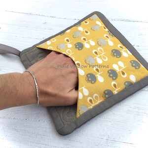May include: A gray and yellow pot holder with a floral pattern and a loop handle. The pot holder has a pocket on the front. "Pixie Willow Patterns" is printed on the front of the pot holder.