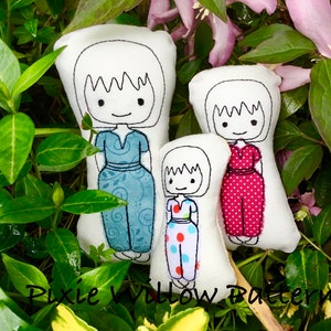 May include: Three white fabric dolls with embroidered faces and outfits. The dolls are dressed in blue, red, and multi-coloured patterned dresses. The dolls are surrounded by green foliage and pink flowers. The text "Pixie Willow Patterns" is at the bottom.