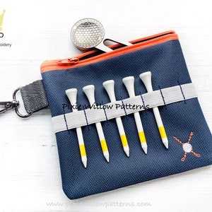 Puede incluir: Un porta-tees de golf azul con cierre de cremallera y una banda elástica blanca para sujetar los tees. El porta-tees tiene una pequeña cremallera naranja y un clip de metal plateado. El porta-tees tiene un pequeño diseño de bola de golf y tee bordado.