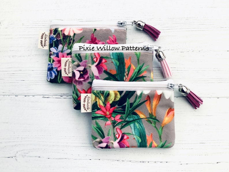 May include: Two small zippered pouches with floral patterns and tassels. The pouches are made of a soft, gray fabric with a white zipper. The pouches are labeled "Handmade with love".