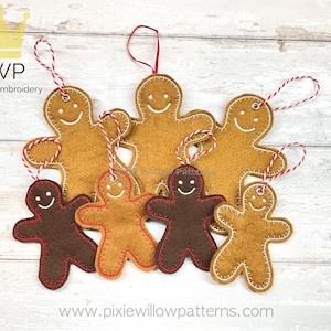 May include: Six felt gingerbread people ornaments in various sizes, with brown and tan felt, red stitching, and white smiling faces. The ornaments are hanging from red and white twine on a light wood background.