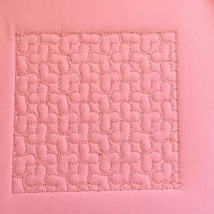 Pode incluir: Um quadrado de tecido rosa com um padrão acolchoado de pequenas formas curvas. O acolchoado é feito com um fio rosa claro.