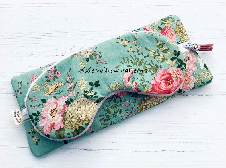 May include: A teal floral eye mask with a white trim and a white fluffy lining. The eye mask is folded in half and has a small silver charm attached to it. The text "Pixie Willow Patterns" is printed on the eye mask.
